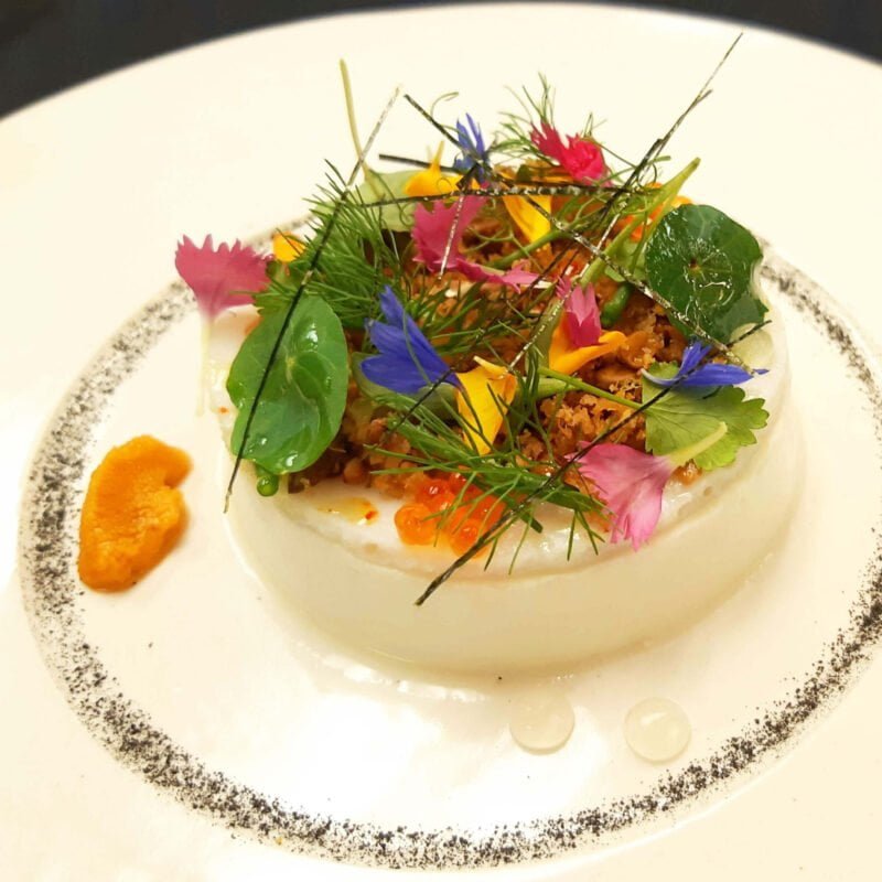 Mousse de coquillages à la mye, le couteau de mer, l'oursin et le buccin. Garni d'herbes aromatiques, fleurs comestibles et algues. Entourée d'une gonade et de deux perles liquides.