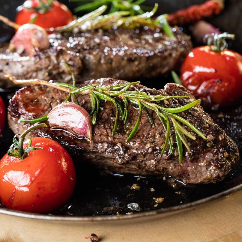 Pièce de steak de phoque frotté au cacao et aux épices accompagné d'ail, tomates et romarin dans une assiette creuse foncée.