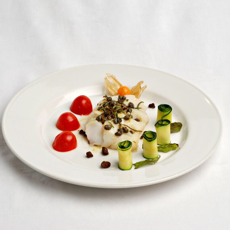 Aiglefin au zeste de citron vert et aux câpres, avec des tomates cerises coupées en deux et des courgettes roulées et posées verticalement et un physalis au fond de l'assiette.