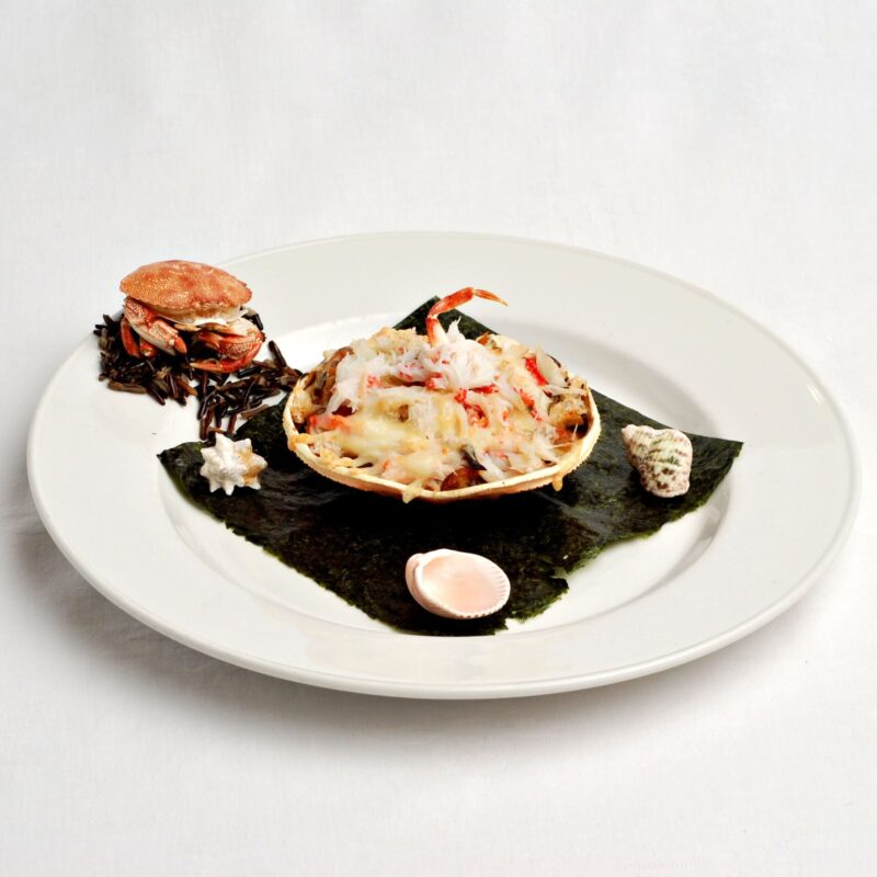 Gratin de crabe dans sa carapace avec des bouts de crabes qui garnissent le dessus on y voit une patte. sur une algue dans une assiette. Avec des coquillage qui décorent et un petit crabe entier sur le haut gauche de l'assiette.