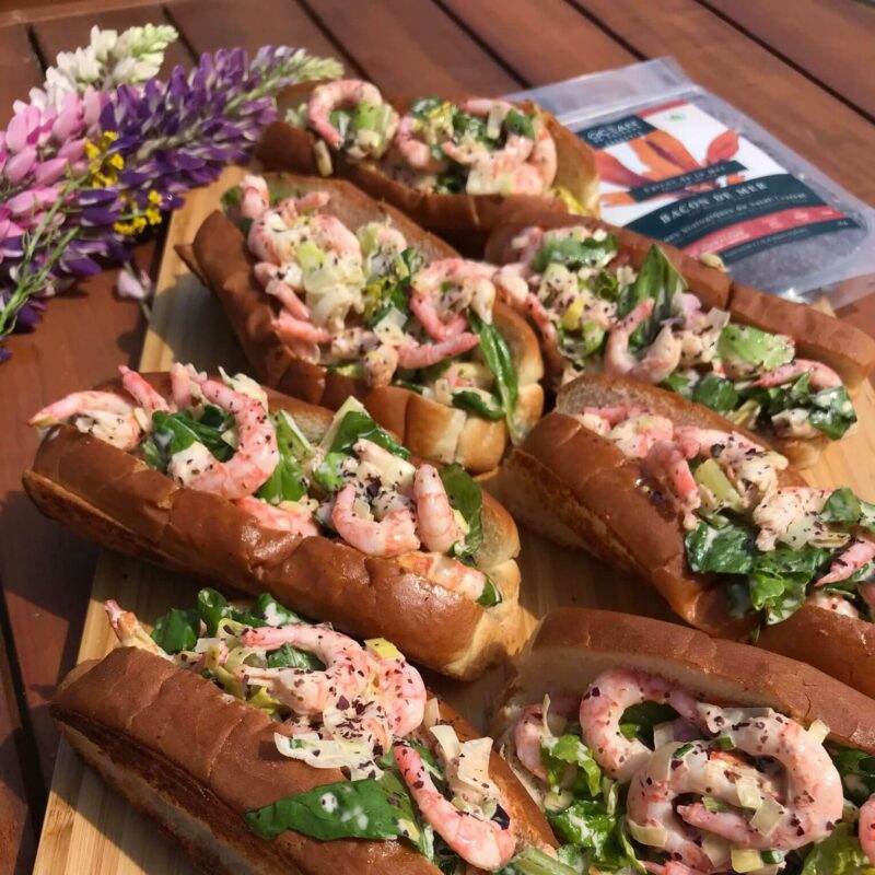 Guédille aux crevettes, bacon de mer, oignons et salade sur une planche en bois.