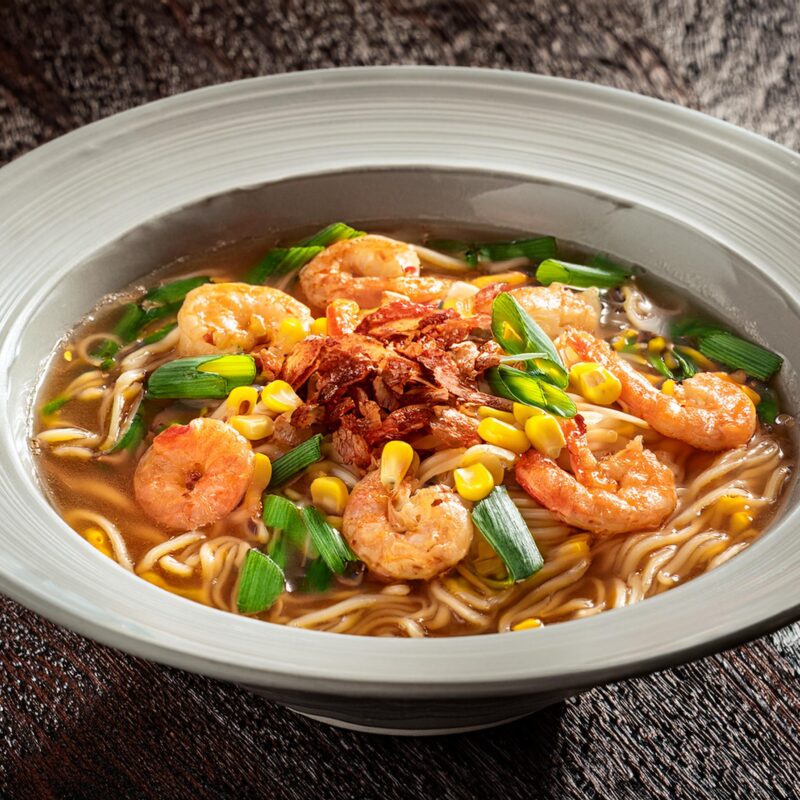 Photographie d'une soupe de nouille aux crevettes avec du maïs, du bacon et des morceaux d'oignons verts en décoration.
