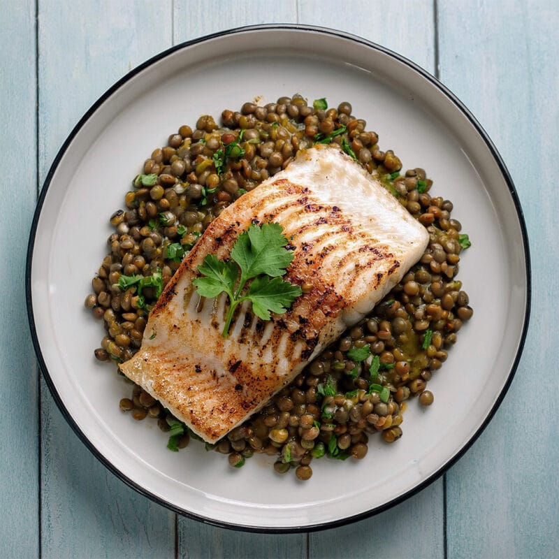 Photographie d'un filet de poisson grillé, posé sur une assiette de lentilles, avec de la coriandre par-dessus.