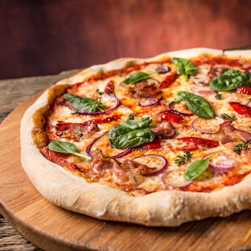 Photographie d'une pizza garnie de bacon, poivrons et tomates, feuilles de basilic.