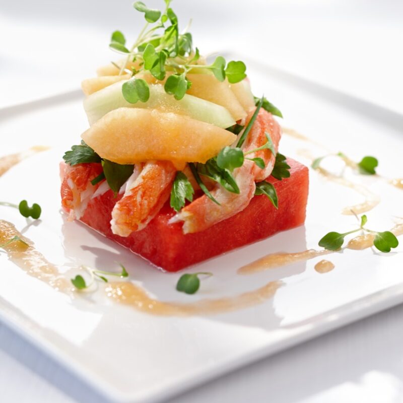 Photographie d'un carré de melon d'eau surmonté de morceaux de crabe avec des morceaux de melon et des petites herbes par-dessus.