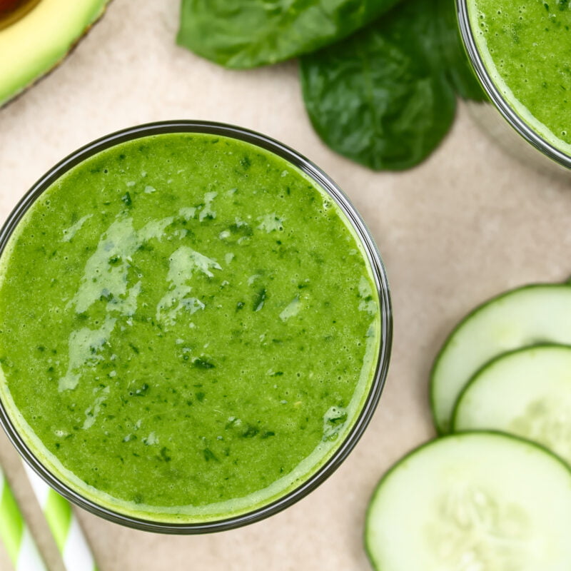 Photographie d'un verre de smoothie couleur verte, prise du dessus. Des rondelles de concombre et un demi avocat sont disposés autour du verre.