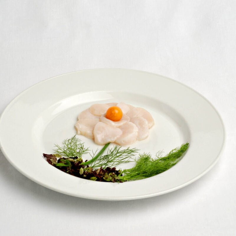 Rosace de pétoncles géants à l’huile de noix et citron vert avec un physalis au milieux et des herbes aromatiques sur le bas de l'assiette.