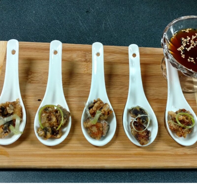 Photographie de 5 petites cuillères avec des morceaux fris dedans et une sauce brun translucide à côté.