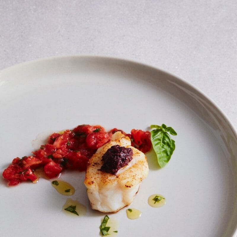 Photographie d'un petit morceau de poisson blanc avec une olive noir par-dessus. À côté se trouve une préparation à base de tomate décorée d'une feuille de basilic.